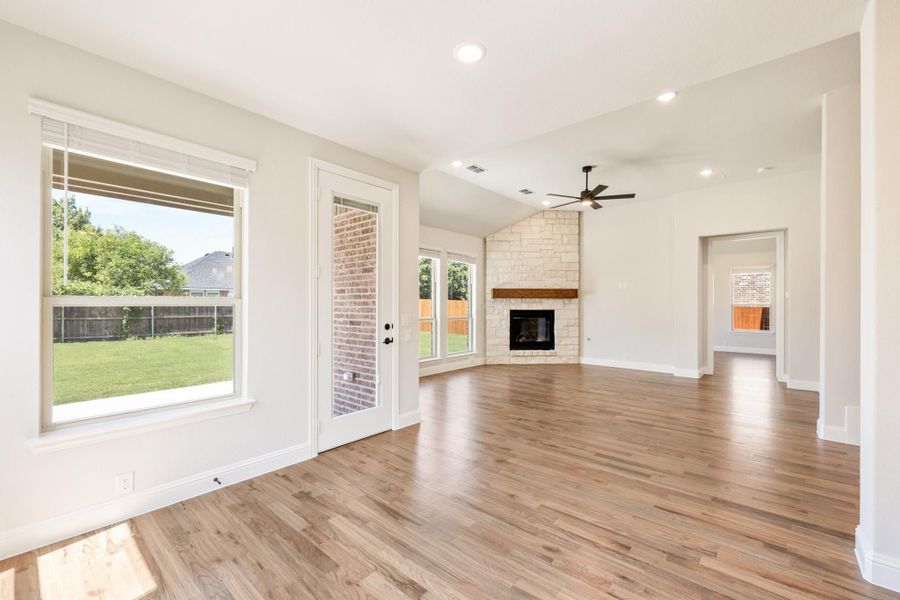 Open living room with hardwood floors, stone corner fireplace, ceiling fan, and backyard views through large windows. Open living room with hardwood floors, stone corner fireplace, ceiling fan, and backyard views through large windows.