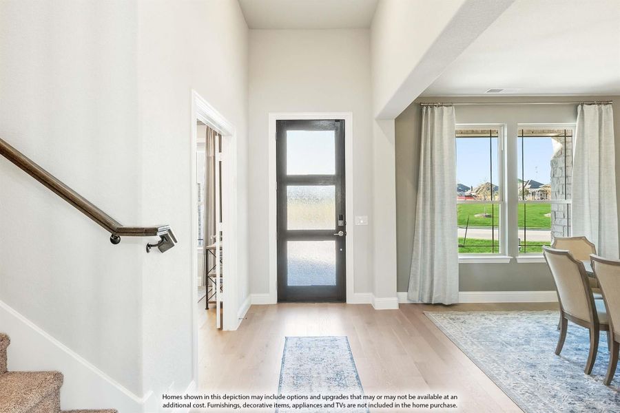 Bright home entryway with glass-panel front door, hardwood floors, staircase, and large windows with curtains.