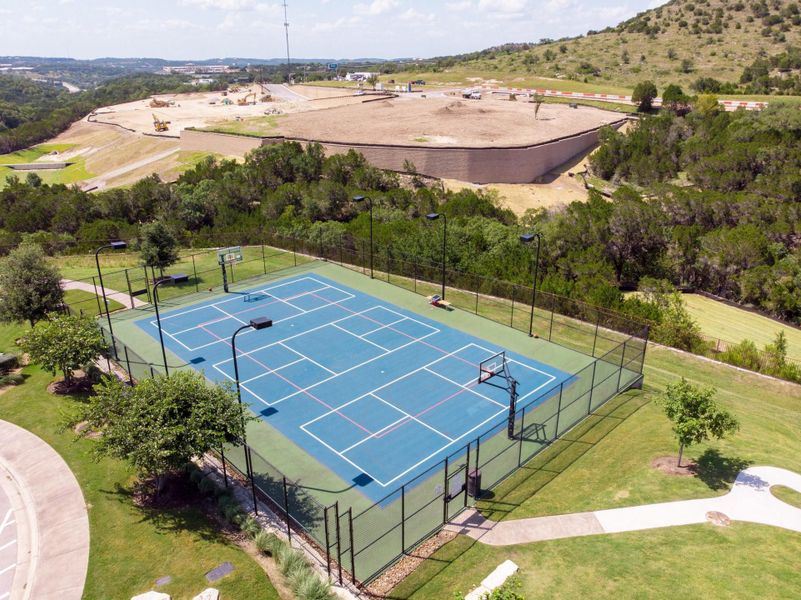 An aerial view showcases a vibrant blue multi-sport court, bordered by lush landscaping and a winding path.