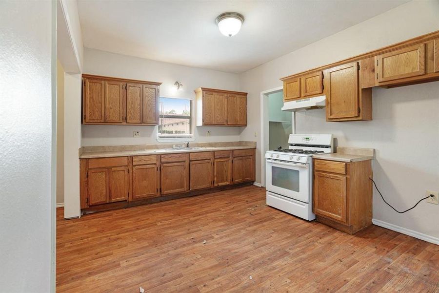 Kitchen with white gas range, light countertops, light wood-style floors, brown cabinetry, and under cabinet range hood Kitchen with white gas range, light countertops, light wood-style floors, brown cabinetry, and under cabinet range hood