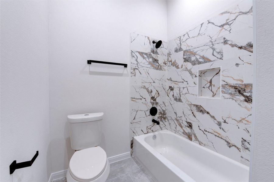 Tub/shower combo finished with marble-look tile featuring bold veining, paired with matte black fixtures for a clean, modern contrast. Built-in niche adds practical storage, while light tile flooring keeps the space bright and cohesive
