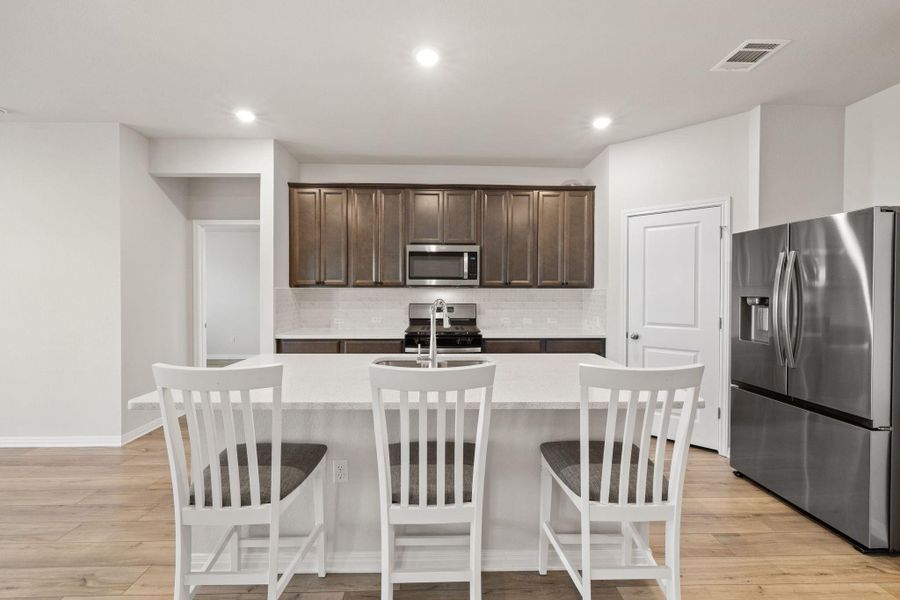 Kitchen featuring stainless steel appliances, dark brown cabinets, a center island with sink, backsplash, and light stone counters Kitchen featuring stainless steel appliances, dark brown cabinets, a center island with sink, backsplash, and light stone counters