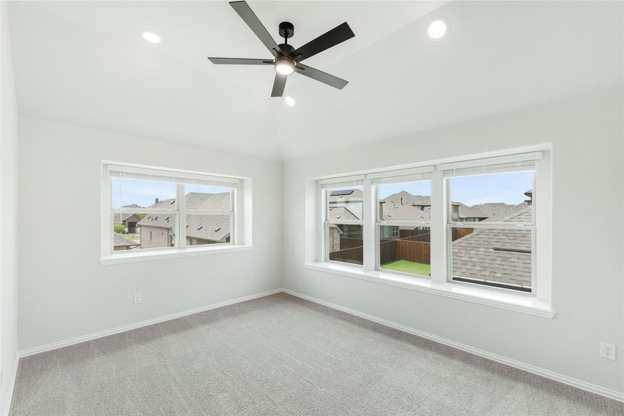 Empty bedroom with gray carpet, ceiling fan, and multiple windows overlooking neighborhood homes