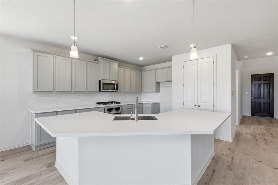 Modern kitchen featuring a large central island with an undermount sink, light gray cabinetry, stainless steel appliances, a white subway tile backsplash, and wood-finish flooring
