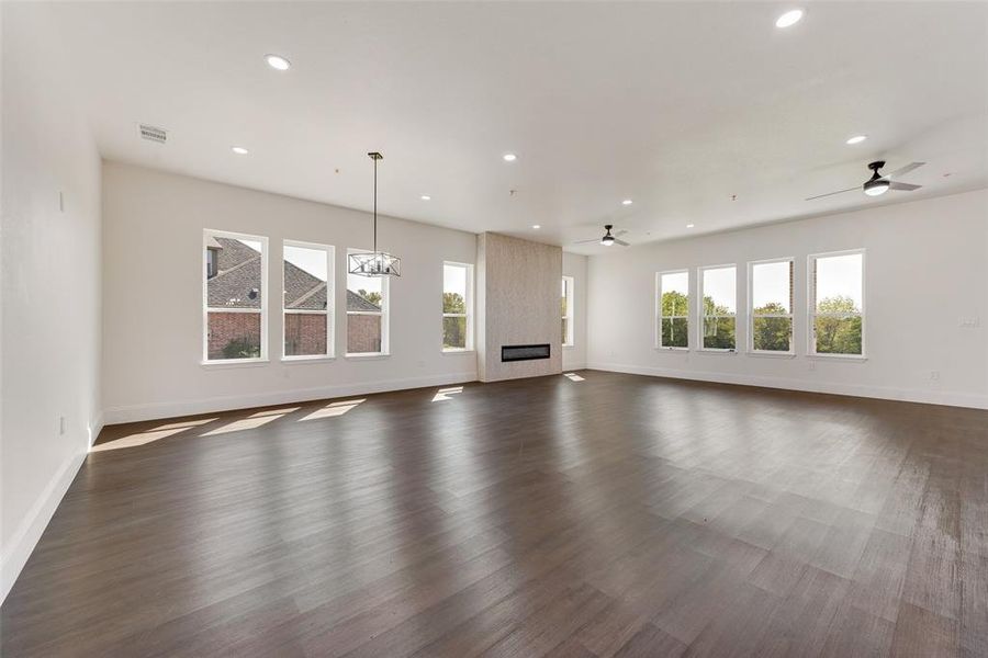 Expansive living area featuring wood-finish flooring, multiple large windows, a contemporary fireplace with a textured surround, recessed lighting, and ceiling fans Expansive living area featuring wood-finish flooring, multiple large windows, a contemporary fireplace with a textured surround, recessed lighting, and ceiling fans