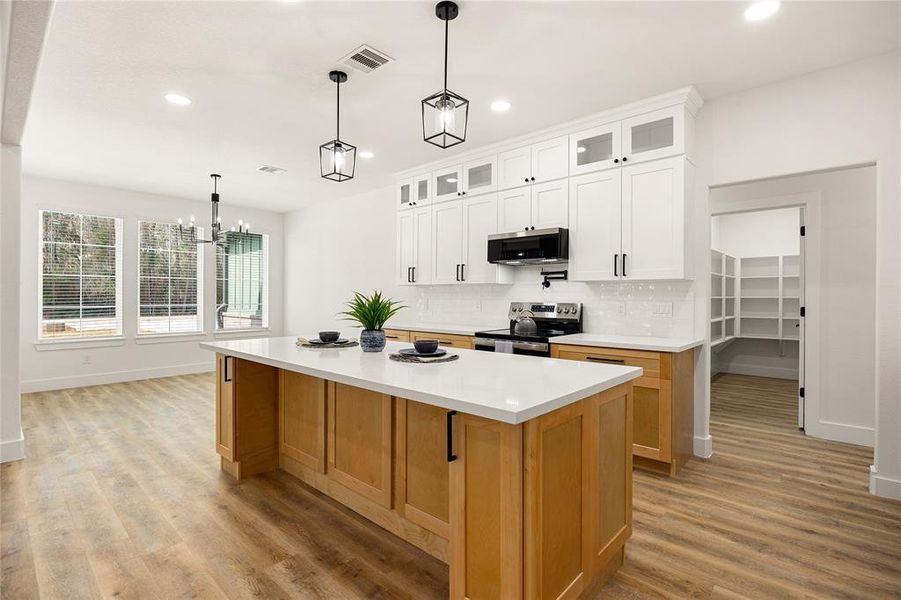 This modern kitchen features a large island with wood accents, sleek white cabinetry, and stainless steel appliances. It's bright and airy with wood flooring and ample natural light from the nearby windows. A pantry is accessible through a side door, offering extra storage.