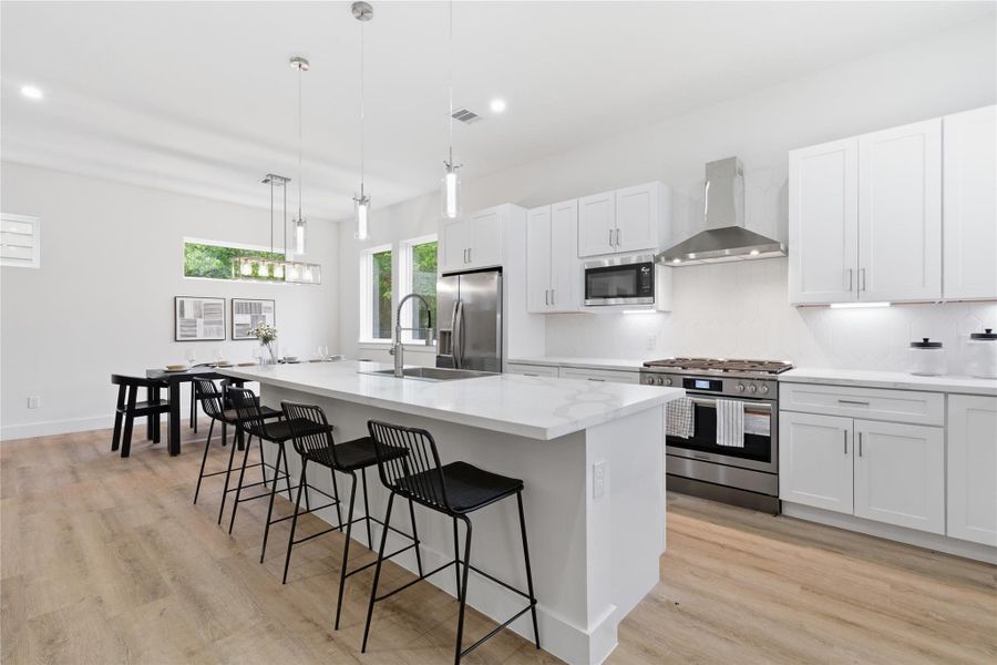 Modern kitchen features sleek white cabinetry, stainless steel appliances, and a large island with bar seating. The open layout connects to a dining area with abundant natural light from large windows, creating a bright and inviting space.