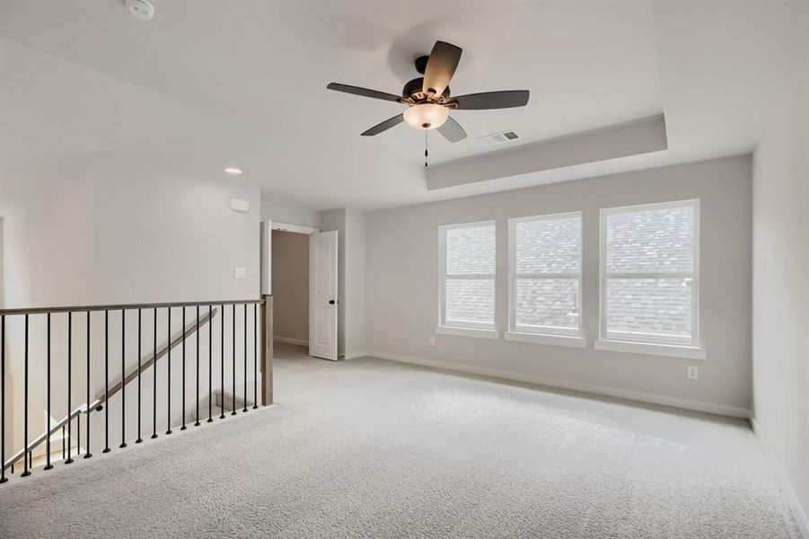 Loft space featuring light gray carpeting, three prominent windows, a ceiling fan with integrated lighting, and a tray ceiling detail
