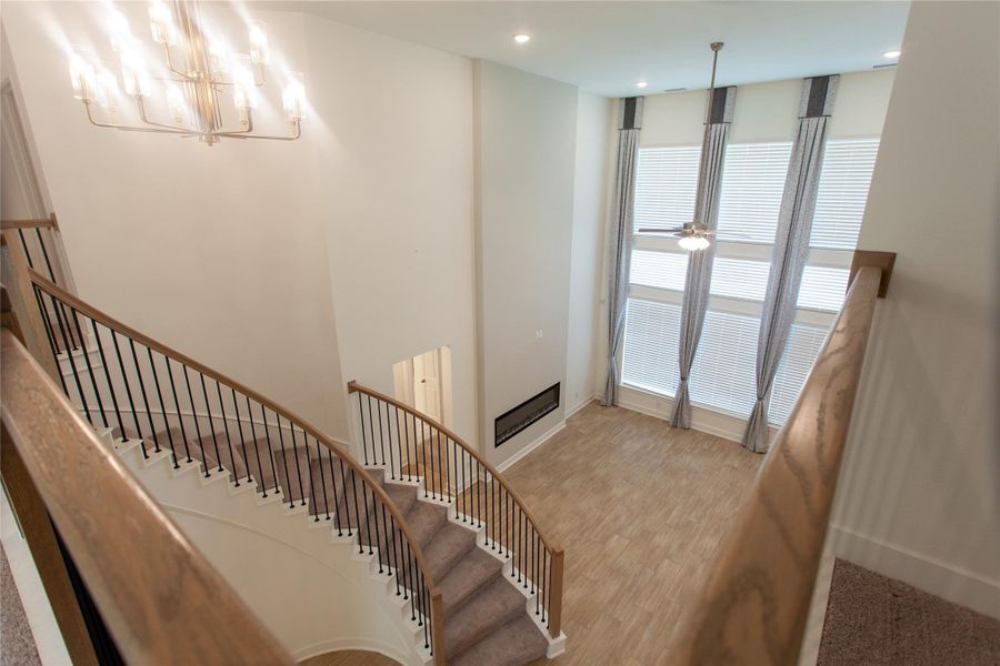 A breathtaking view from the upstairs landing showcasing the curved staircase, dramatic chandelier, two-story window wall, custom drapery, ceiling fan, and built-in linear fireplace below — every angle of this home tells a story.