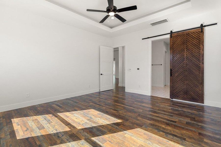 Unfurnished bedroom featuring a barn door, a raised ceiling, ceiling fan, and dark wood finished floors