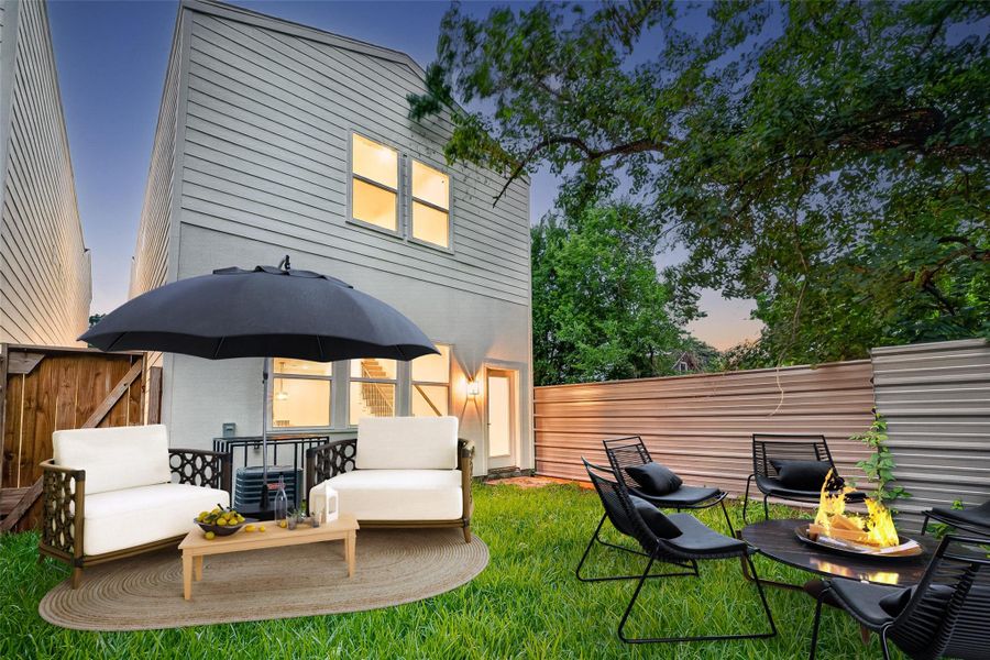 This photo showcases a modern backyard with a cozy seating area featuring white chairs, a large umbrella, and a fire pit. The space is surrounded by a privacy fence and lush greenery, providing an inviting atmosphere for relaxation and entertaining.- Virtual Staging This photo showcases a modern backyard with a cozy seating area featuring white chairs, a large umbrella, and a fire pit. The space is surrounded by a privacy fence and lush greenery, providing an inviting atmosphere for relaxation and entertaining.- Virtual Staging