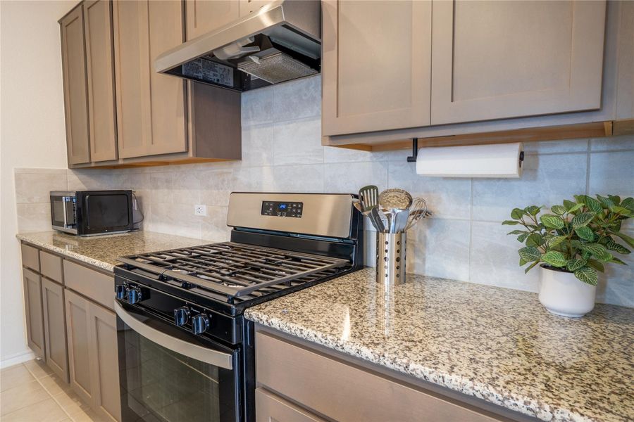 Modern kitchen featuring a stainless steel range with overhead ventilation, granite countertops, tile backsplash, and neutral cabinetry Modern kitchen featuring a stainless steel range with overhead ventilation, granite countertops, tile backsplash, and neutral cabinetry