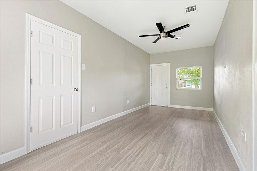 This room features a clean, modern design with light-colored wood flooring and neutral walls. It includes a ceiling fan, a window for natural light, and a white door. Perfect for a bedroom or office space.