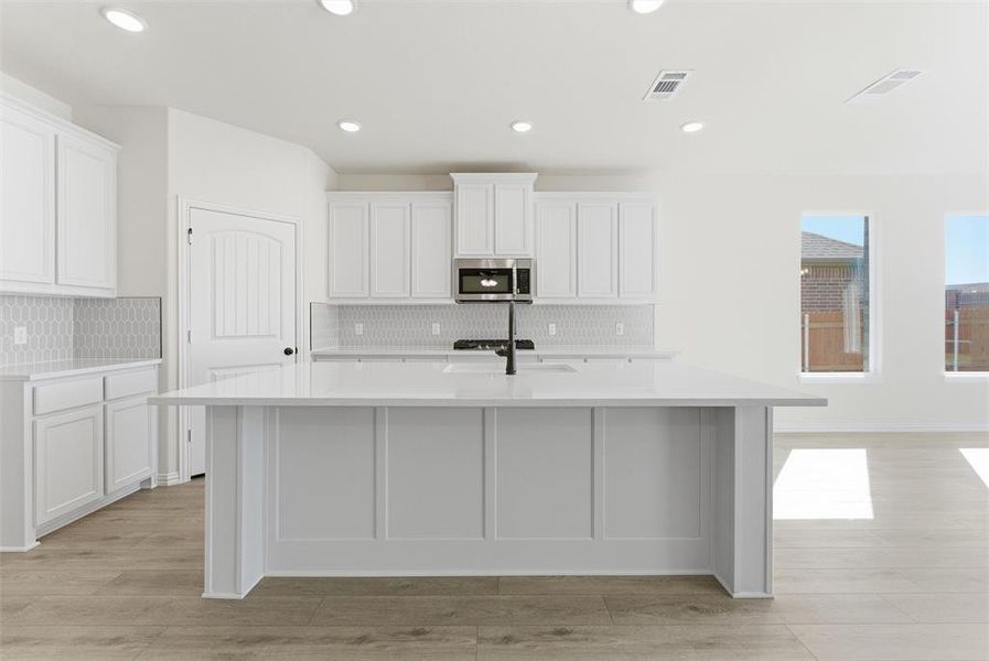 Kitchen featuring white cabinetry, recessed lighting, stainless steel microwave, light stone countertops, and an island with sink