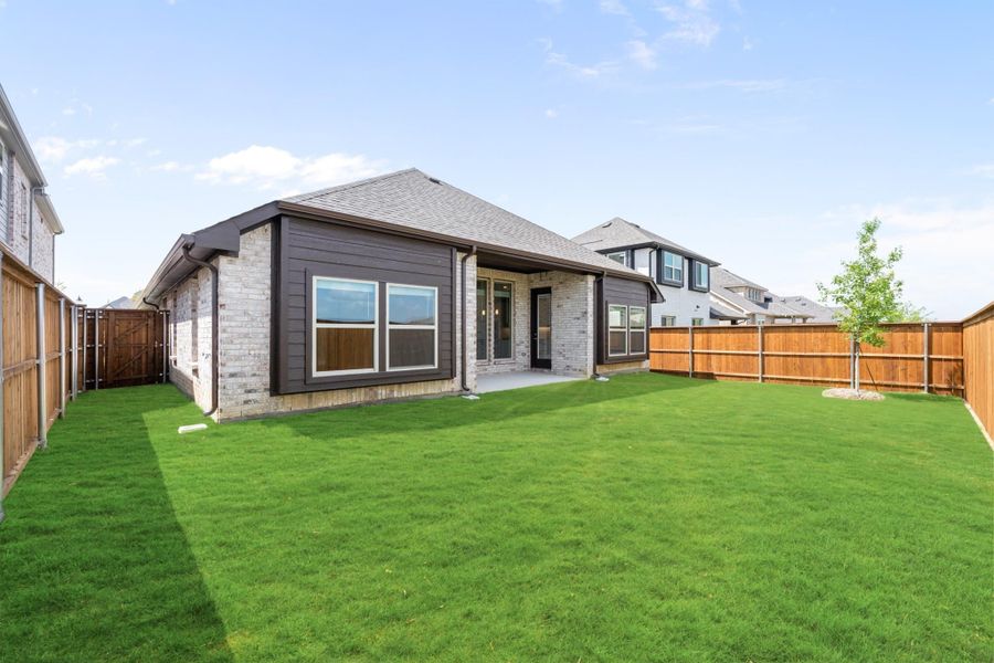 Rear view of single-story brick home with dark trim, covered patio, and fenced backyard with green lawn Rear view of single-story brick home with dark trim, covered patio, and fenced backyard with green lawn