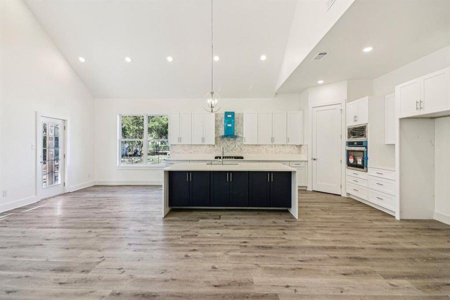 Kitchen featuring white cabinets, a kitchen island with sink, decorative backsplash, high vaulted ceiling, and decorative light fixtures Kitchen featuring white cabinets, a kitchen island with sink, decorative backsplash, high vaulted ceiling, and decorative light fixtures