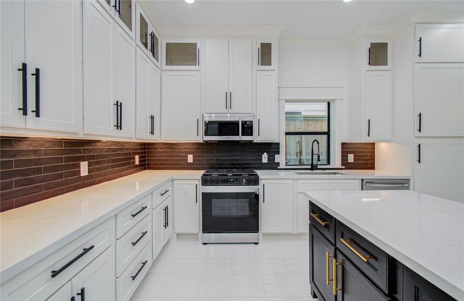Kitchen features sleek white cabinetry with black hardware, a stylish dark tile backsplash, and stainless steel appliances. The spacious layout includes a central island with a contrasting dark cabinetry and gold accents, ideal for cooking and entertaining.