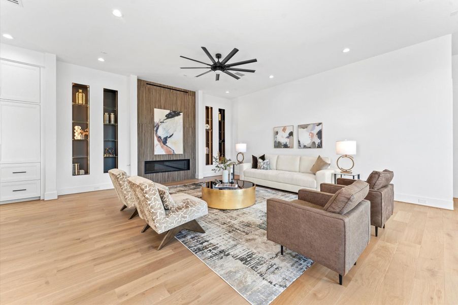 This spacious living room features a modern design with a sleek fireplace, built-in shelving, and recessed lighting. It includes comfortable seating, stylish decor, and a large ceiling fan, all set on elegant hardwood floors.