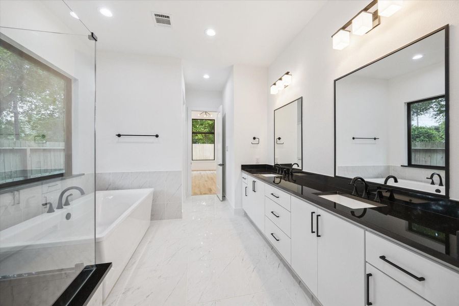 The spa-inspired primary bath features a freestanding soaking tub, spacious glass-enclosed shower with a built-in bench, and dual vanities with sleek black countertops and framed mirrors. Large-format marble-look tile adds a polished touch, while the black fixtures and hardware bring bold contrast and timeless appeal. The spa-inspired primary bath features a freestanding soaking tub, spacious glass-enclosed shower with a built-in bench, and dual vanities with sleek black countertops and framed mirrors. Large-format marble-look tile adds a polished touch, while the black fixtures and hardware bring bold contrast and timeless appeal.