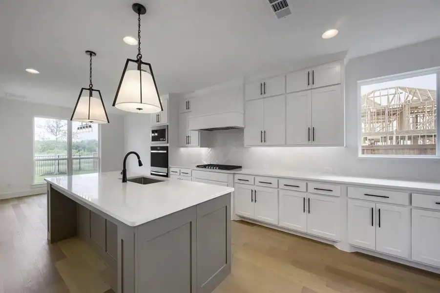 Bright kitchen featuring a central island with a sink and matte black faucet, pendant lighting, white cabinetry with dark hardware, and light wood-finish flooring Bright kitchen featuring a central island with a sink and matte black faucet, pendant lighting, white cabinetry with dark hardware, and light wood-finish flooring