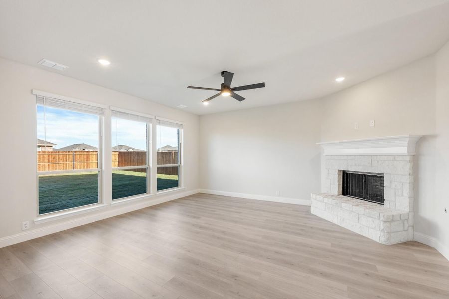 Open living room with light wood floors, stone fireplace, ceiling fan, and large windows overlooking backyard Open living room with light wood floors, stone fireplace, ceiling fan, and large windows overlooking backyard
