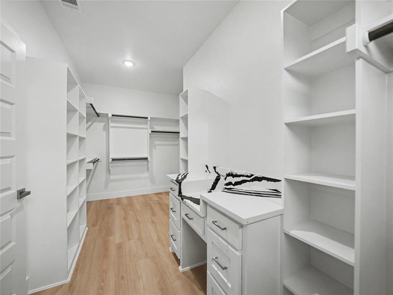 Spacious walk-in closet featuring light wood-finish flooring, recessed lighting, extensive white shelving, multiple clothing rods, and a built-in white vanity with drawers Spacious walk-in closet featuring light wood-finish flooring, recessed lighting, extensive white shelving, multiple clothing rods, and a built-in white vanity with drawers