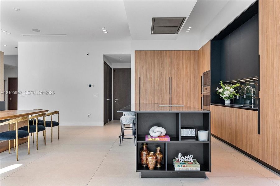 Modern kitchen featuring wood-finish cabinetry, matte black integrated shelving, a central island with storage, built-in wall appliances, and tile flooring