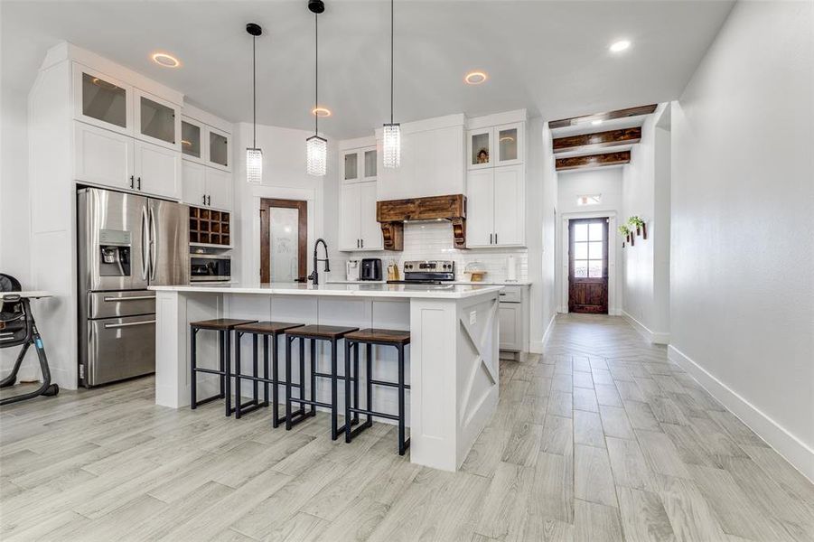 Kitchen featuring light countertops, appliances with stainless steel finishes, tasteful backsplash, an island with sink, and glass insert cabinets