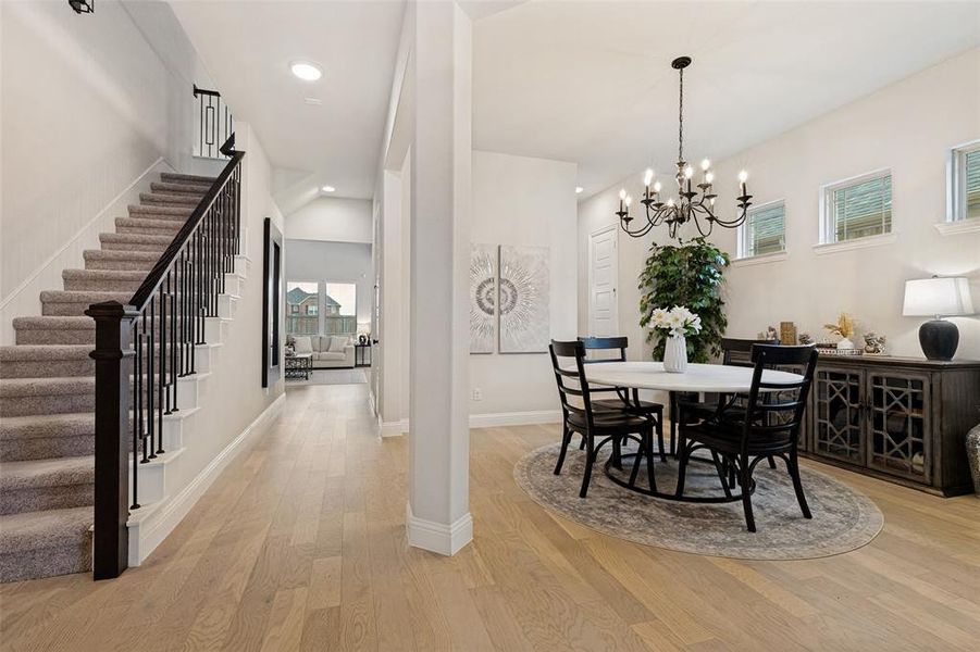 Open-concept living space featuring wood-finish flooring, a carpeted staircase with dark iron balusters, a formal dining area with a chandelier, and ample natural light from multiple windows
