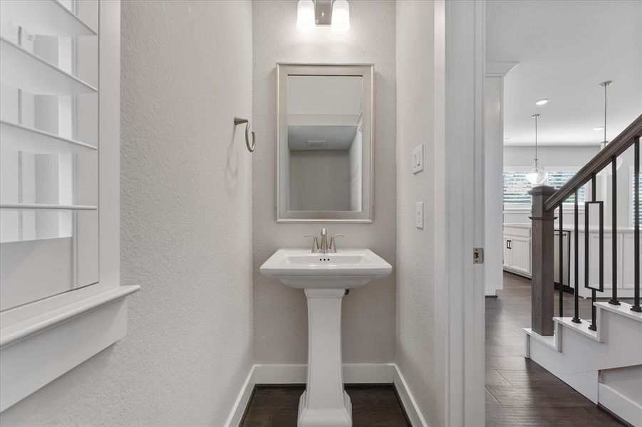 This  well-lit powder room featuring a pedestal sink, a framed mirror, and built-in shelving, situated conveniently adjacent to a staircase and main living areas.