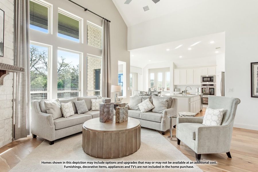 Open-concept living room with vaulted ceiling, tall windows, stone fireplace, and kitchen visible in background