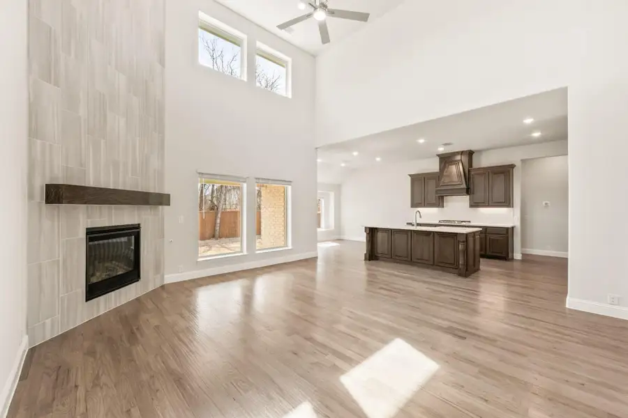 Open living area with two-story ceiling, tiled fireplace, hardwood floors, and kitchen with dark cabinets visible in background