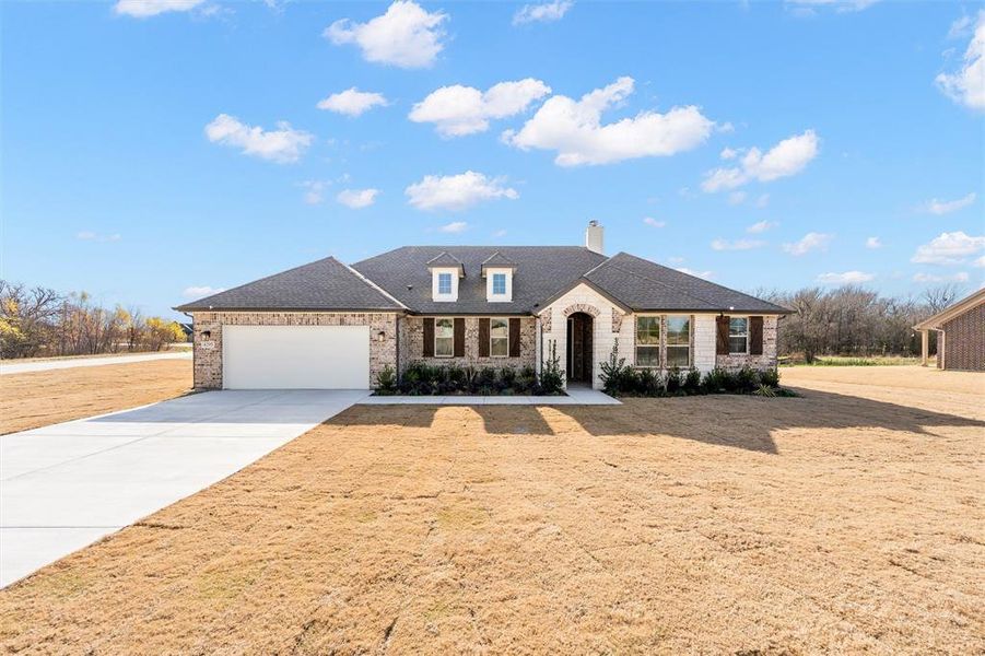 French country inspired facade with a chimney, concrete driveway, brick siding, an attached garage, and roof with shingles