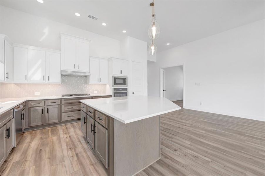 Contemporary kitchen featuring a large central island with a solid surface countertop, two-tone cabinetry, stainless steel appliances, a tiled backsplash, and recessed lighting Contemporary kitchen featuring a large central island with a solid surface countertop, two-tone cabinetry, stainless steel appliances, a tiled backsplash, and recessed lighting