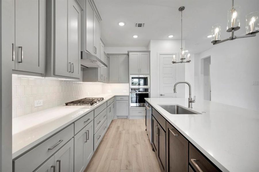 Kitchen featuring stainless steel appliances, light wood-type flooring, suspended lighting, tasteful backsplash, and light stone counters Kitchen featuring stainless steel appliances, light wood-type flooring, suspended lighting, tasteful backsplash, and light stone counters