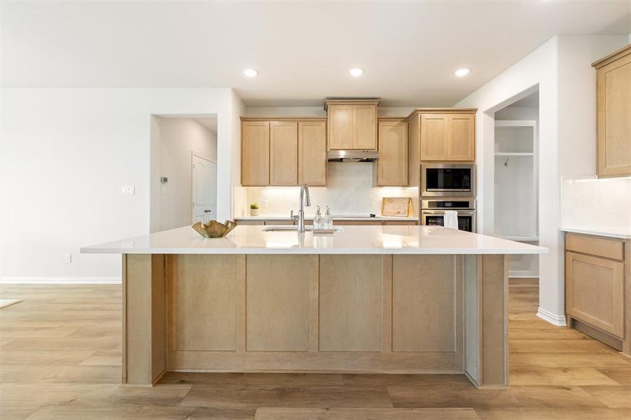 Kitchen with light brown cabinetry, appliances with stainless steel finishes, a kitchen island with sink, light stone countertops, and recessed lighting