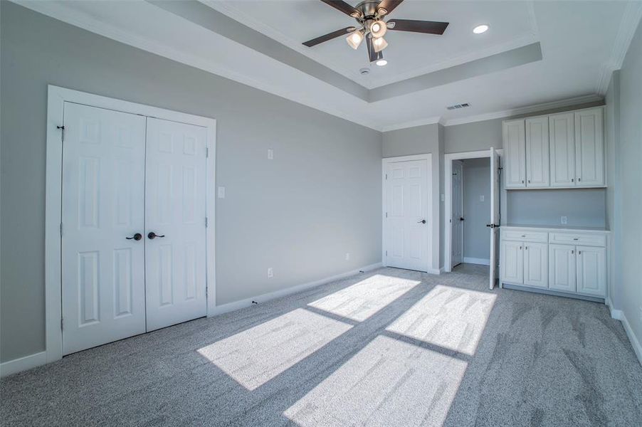 Unfurnished bedroom with light colored carpet, a ceiling fan, a raised ceiling, crown molding, and recessed lighting Unfurnished bedroom with light colored carpet, a ceiling fan, a raised ceiling, crown molding, and recessed lighting