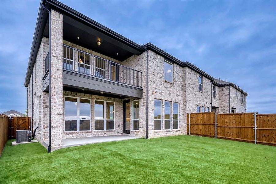 Brick exterior with a covered patio, second-story balcony with metal railing, and a fenced yard with artificial turf