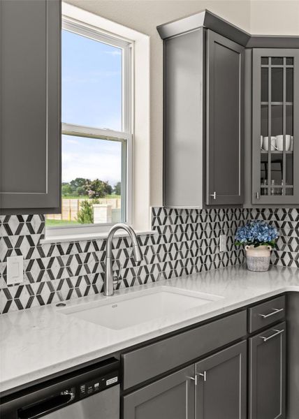 Kitchen with gray cabinets, stainless steel dishwasher, glass fronted cabinets, and light stone counters
