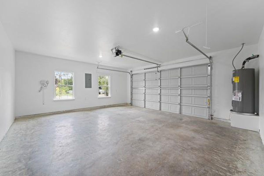 Spacious garage featuring a concrete floor, bright white walls, two garage doors with automatic openers, two windows providing natural illumination, and a visible water heater Spacious garage featuring a concrete floor, bright white walls, two garage doors with automatic openers, two windows providing natural illumination, and a visible water heater