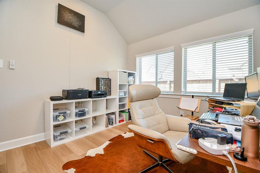 This photo shows a bright home office with large windows, a modern office chair, a desk with electronics, and organized shelving. The space has a cozy rug and light hardwood floors, creating a comfortable and functional work environment.