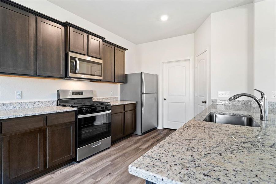 Kitchen with light stone countertops, dark brown cabinets, light wood-type flooring, sink, and appliances with stainless steel finishes