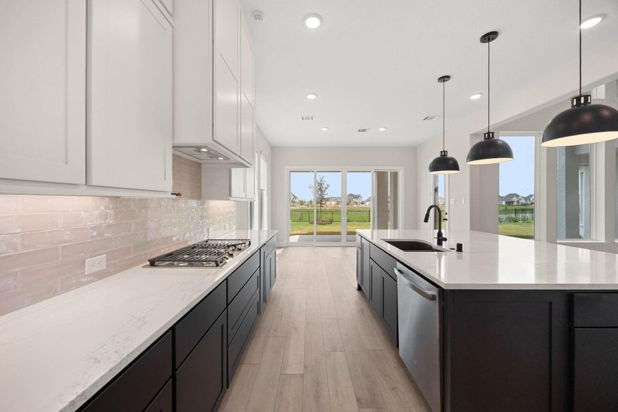 Detailed view of the premium culinary zone, highlighting the sleek gas cooktop and long quartz countertops. This angle showcases the beautiful, glossy tile backsplash that catches the light under the upper cabinets, alongside the highly functional galley-style layout. The expansive island houses a stainless steel dishwasher and an undermount sink, perfectly matching the modern matte black pendant lights and offering a clear view toward the scenic backyard. Detailed view of the premium culinary zone, highlighting the sleek gas cooktop and long quartz countertops. This angle showcases the beautiful, glossy tile backsplash that catches the light under the upper cabinets, alongside the highly functional galley-style layout. The expansive island houses a stainless steel dishwasher and an undermount sink, perfectly matching the modern matte black pendant lights and offering a clear view toward the scenic backyard.