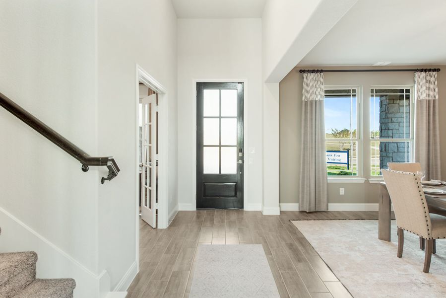 Bright home entryway with dark front door, wood-look floors, staircase, and adjacent dining area with large windows.