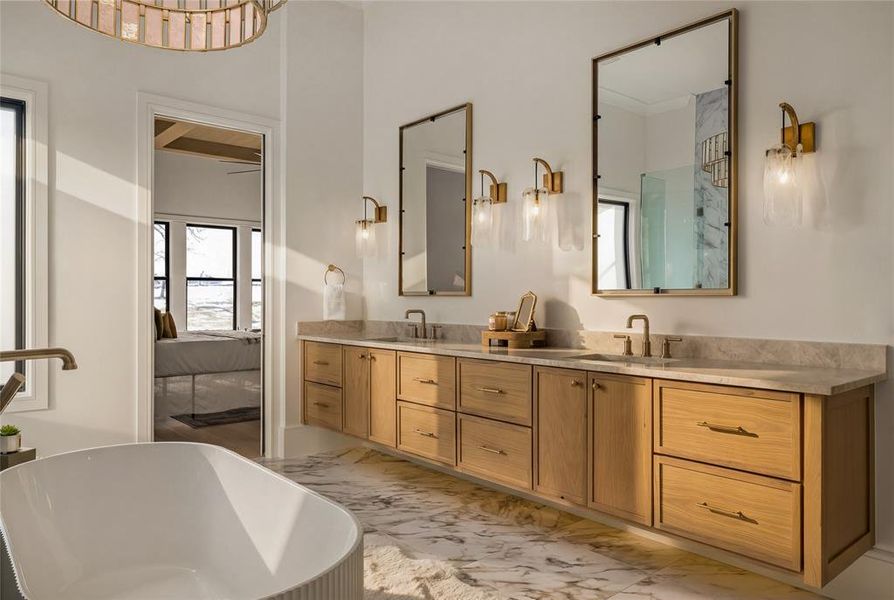 Bathroom featuring a freestanding soaking tub, a dual vanity with light wood cabinetry, stone countertops, and rectangular brass-framed mirrors