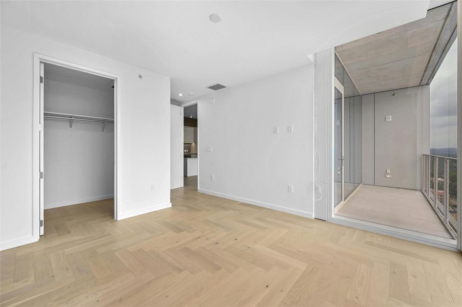 The spacious primary bedroom features elegant light wood herringbone flooring and clean, modern lines throughout. Floor-to-ceiling window allowing for abundant natural light. The room includes a generous walk-in closet with built-in shelving and hanging space.