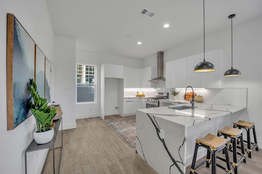 Kitchen featuring pendant lighting, kitchen peninsula, stainless steel gas stove, wall chimney range hood, and white cabinetry