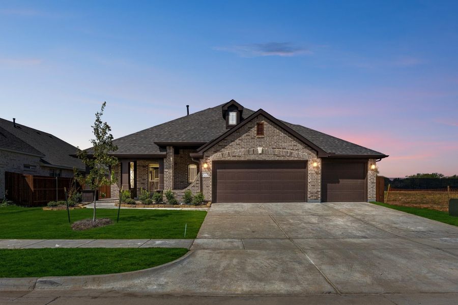 Single-story brick home with two-car garage, dark roof, and landscaped front yard at dusk