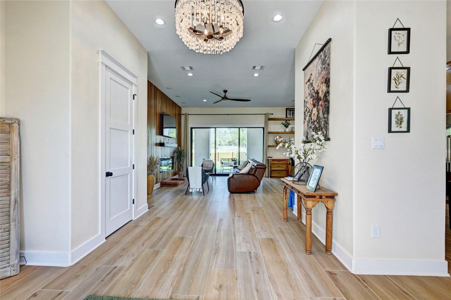 This photo showcases a bright, open-concept living area with light wood flooring and neutral walls. A modern chandelier and ceiling fan add elegance and comfort. The space connects to a cozy seating area with a view of greenery through large glass doors, enhancing natural light.