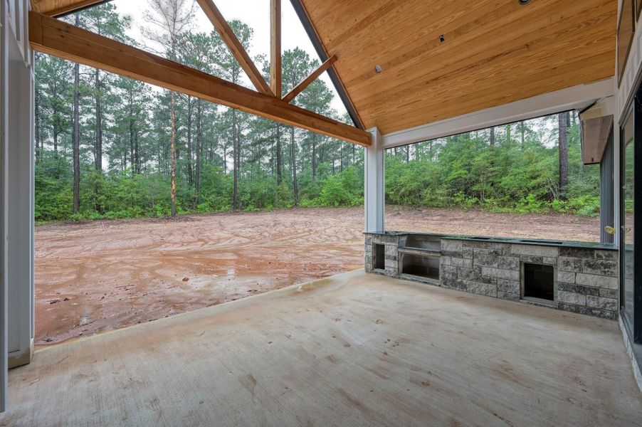 The spacious covered patio with a vaulted wooden ceiling, stone accents, and an outdoor kitchen area. It overlooks a large, open yard with a backdrop of lush trees, providing a serene and private setting.